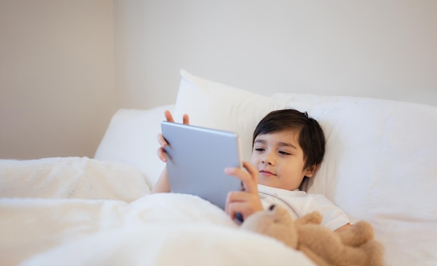 Un enfant heureux allongé dans son lit tenant une tablette regardant des dessins animés et discutant avec ses amis sur un bloc-notes numérique Un jeune garçon mignon jouant à des jeux en ligne sur Internet Un enfant se détendant le matin avant d'aller à l'école