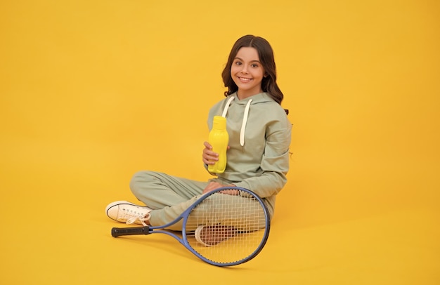 Enfant heureux assis dans des vêtements de sport avec une bouteille d'eau de raquette de tennis sur l'équilibre hydrique de fond jaune