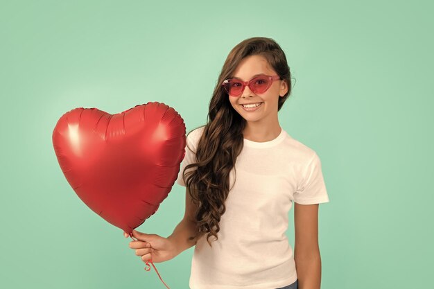 Un enfant heureux dans des lunettes de soleil tient un ballon de fête coeur rouge pour la saint valentin symbole d'amour chérie