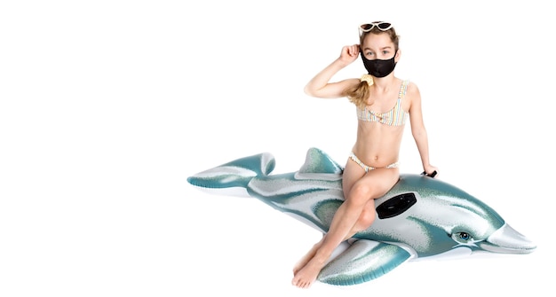 Enfant heureux dans un masque médical noir covid19 en maillot de bain sur un dauphin gonflable. Photographie de studio sur fond blanc. Pandémie de coronavirus et concept de mode de vie sain. photo de haute qualité