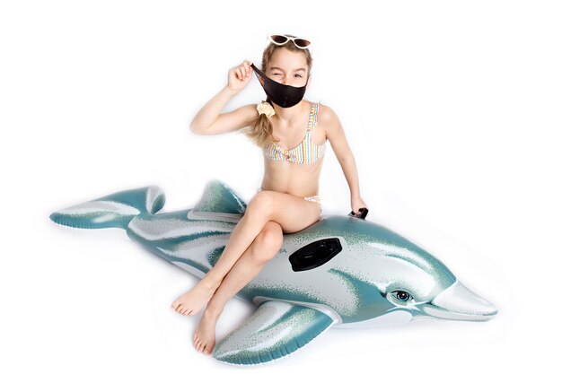 Enfant heureux dans un masque médical noir covid19 en maillot de bain sur un dauphin gonflable. Photographie de studio sur fond blanc. Pandémie de coronavirus et concept de mode de vie sain. photo de haute qualité