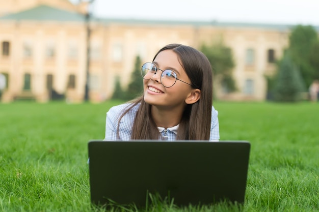 Un enfant heureux dans des verres apprend en ligne dans une école virtuelle à l'aide de la technologie d'un ordinateur portable sur l'herbe verte à l'extérieur