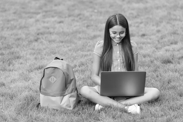 Un enfant heureux et intelligent porte des lunettes à l'aide d'un ordinateur portable pour faire ses devoirs scolaires en ligne assis dans un parc sur l'herbe verte avec un sac à dos