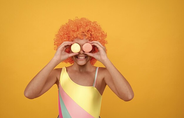 Enfant heureux en maillot de bain portant des cheveux perruque bouclée orange tenant une dent sucrée macaron français