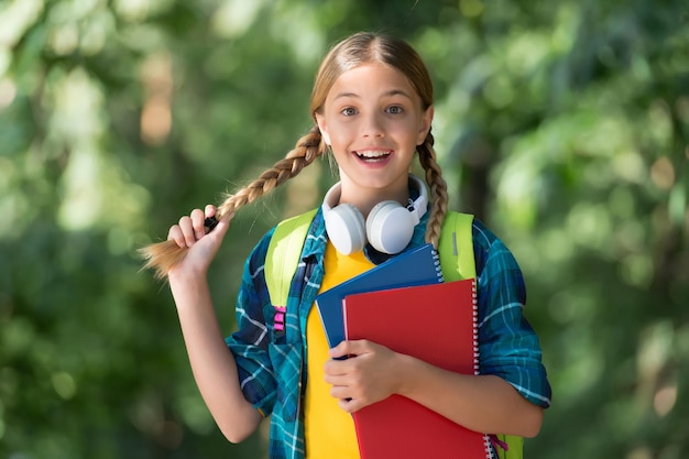 Un enfant heureux avec des nattes porte des livres scolaires pour l'apprentissage des langues étrangères avec des écouteurs paysage naturel, anglais.