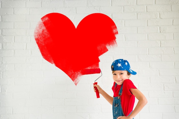 Enfant heureux peignant un grand coeur rouge sur le mur. Fille drôle jouant à la maison. Carte de Saint Valentin. Concept de rénovation et de design