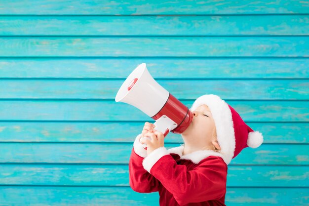 Enfant heureux portant le costume de père Noël parlant par mégaphone. Portrait d'enfant drôle sur fond bleu