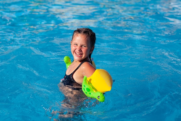 Un enfant heureux pose dans la piscine, une petite fille souriante dans des brassards gonflables avec des dinosaures apprend à nager en p...