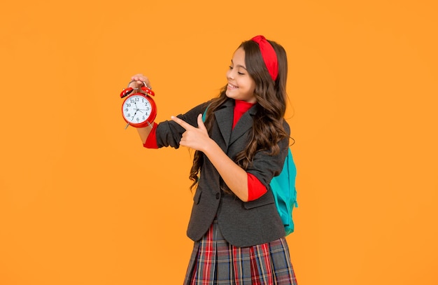 enfant heureux en uniforme scolaire avec sac à dos pointez le doigt sur l'horloge de réveil sur fond jaune matin