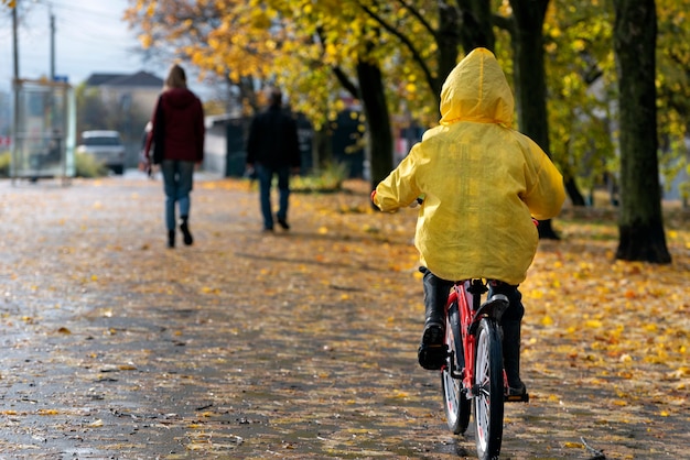 Un enfant en imperméable jaune roule le long de l'allée d'automne avec des gens en arrière-plan. Jour d'automne pluvieux. Vue arrière.