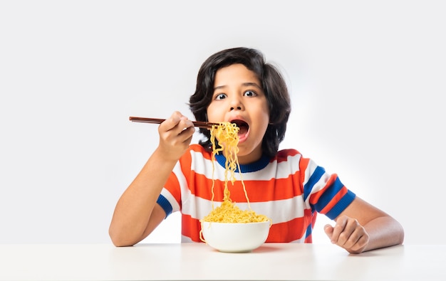 Enfant indien mangeant de délicieuses nouilles avec une fourchette sur fond blanc, un garçon asiatique mange des spaghettis dans un bol sur fond blanc