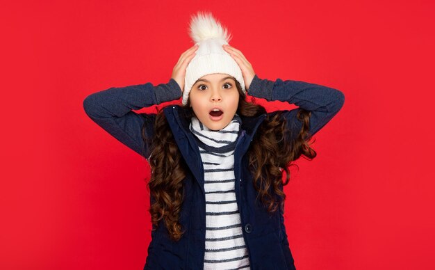 Enfant inquiet choqué avec des cheveux bouclés en chapeau adolescente sur fond rouge