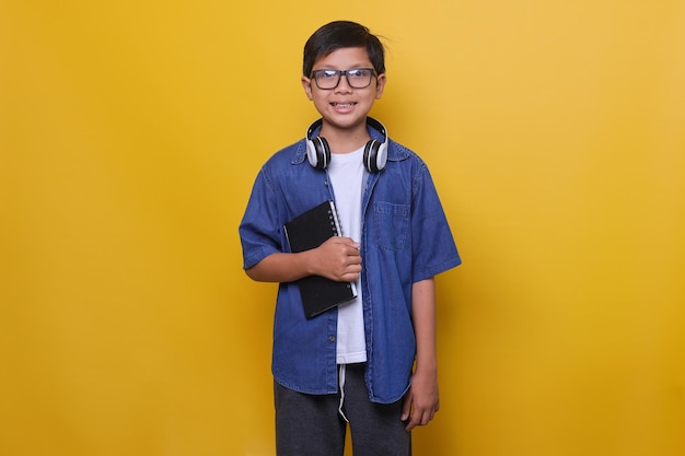 Un enfant intelligent dans des écouteurs et des lunettes de vêtements décontractés est debout et tient un livre souriant au