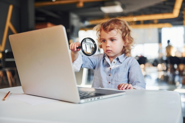 Un enfant intelligent dans des vêtements décontractés avec un ordinateur portable sur la table s'amuse avec une loupe