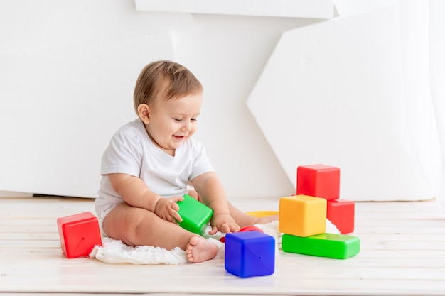 L'enfant joue, heureux petit bébé de six mois dans un t-shirt blanc et des couches jouant à la maison sur un tapis dans une pièce lumineuse avec des cubes de couleurs vives