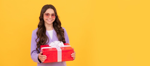 Enfant joyeux à la mode en lunettes de soleil et costume tenir une boîte-cadeau sur fond jaune shopping Fille enfant avec affiche horizontale cadeau En-tête de bannière avec espace de copie