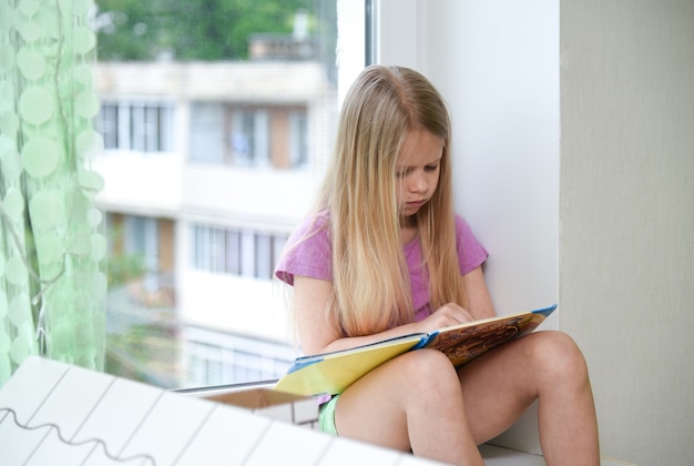 Enfant lisant un livre l'après-midi à la fenêtre.