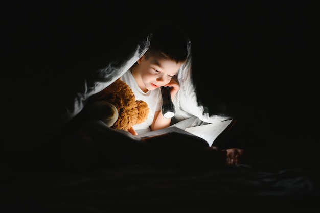 Enfant lisant un livre au lit. Les enfants lisent le soir. Petit garçon avec des livres de contes de fées dans la chambre. L'éducation des jeunes enfants. Histoire au coucher le soir. Enfant mignon sous couverture dans une pièce sombre avec lampe.
