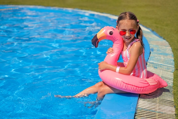 Enfant en maillot de bain rayé et lunettes de soleil est assis au bord de la piscine