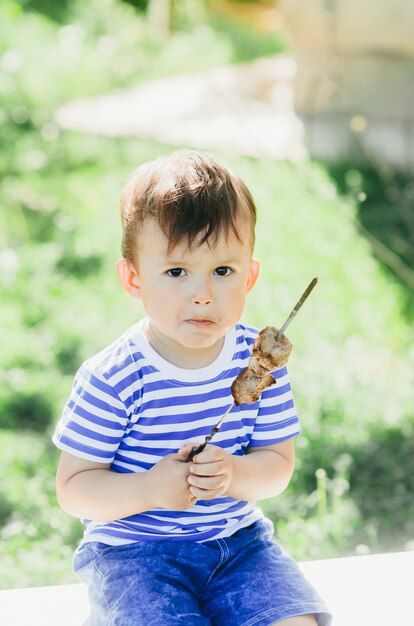 Un enfant mange un kebab sur une brochette, dans un parc ou une forêt nature fond vert d'arbres et d'herbe