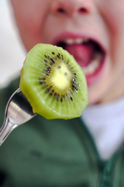 Un enfant mangeant un kiwi avec sa bouche ouverte une tranche de kiwi coincée dans une fourchette