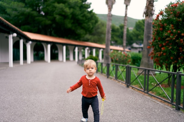 L'enfant marche sur l'asphalte dans la perspective d'un long pavillon dans le parc parmi la verdure