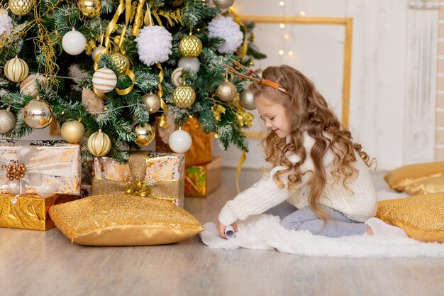 Un enfant met une lettre pour le Père Noël sous un arbre de Noël avec un décor d'or à la maison