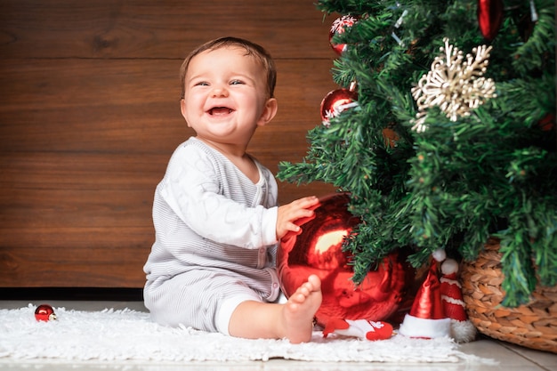 Enfant mignon avec arbre de Noël. bébé heureux assis près d'un sapin et tenant une décoration de Noël et souriant