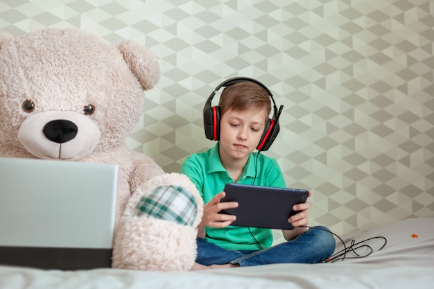 Enfant mignon dans les écouteurs jouant sur une tablette numérique à côté d'un ours en peluche à la recherche dans l'ordinateur à la maison.