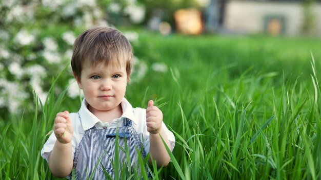 Un enfant mignon dans une salopette bleue et des yeux bleus joue drôlement dans la grande herbe verte dans un parc fleuri vert