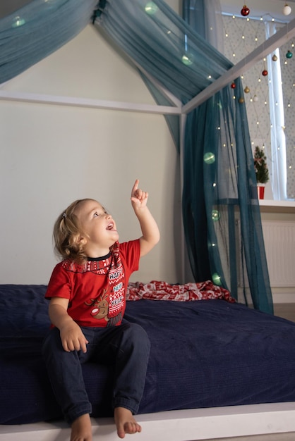 Un enfant mignon impressionné par la décoration de Noël dans sa chambre, la fille est assise dans le lit en t-shirt rouge