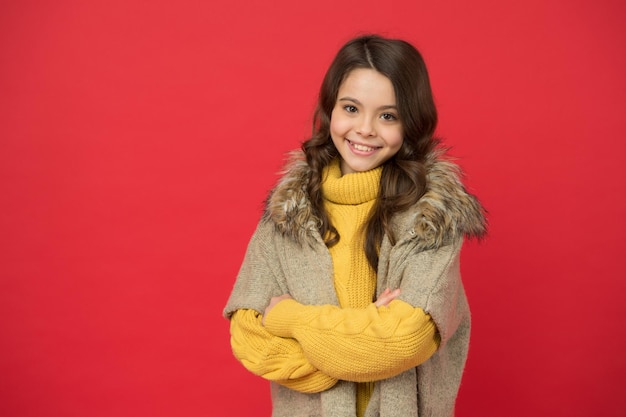 Un enfant mignon souriant a de longs cheveux bouclés portant un pull tricoté chaud et un gilet d'automne sur fond rouge, style de saison.