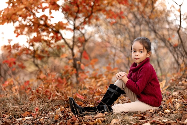 Enfant à la mode de 5 ans posant dans le parc en automne. petite fille assise dans les feuilles tombées