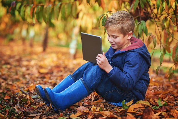 Enfant moderne Photo pleine longueur d'un adorable petit garçon à l'aide d'une tablette assis à l'extérieur pendant l'automne