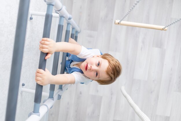 Un enfant sur le mur suédois fait du sport à la maison, un garçon monte une échelle avec une corde, le concept de sport et de santé.