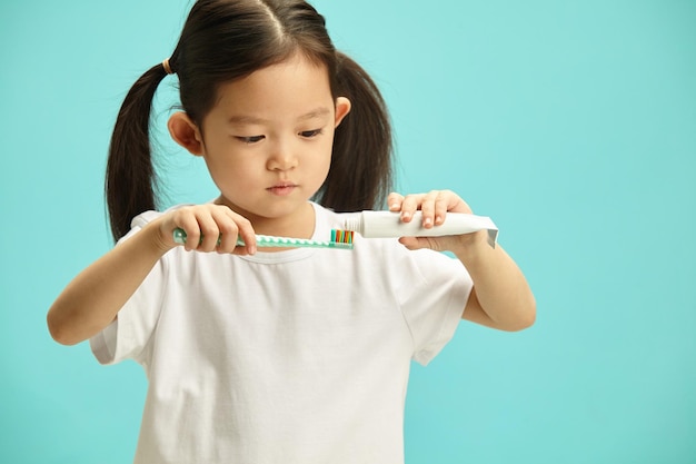 Enfant d'origine asiatique pressant du dentifrice à partir d'un tube sur une brosse à dents debout au-dessus d'un bleu isolé
