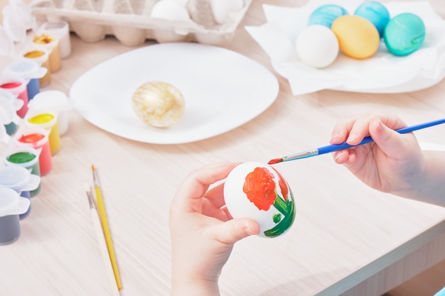 Enfant peint oeuf de Pâques sur une table en bois, boîte d'oeufs, assiette, pinceaux et peintures sur la table