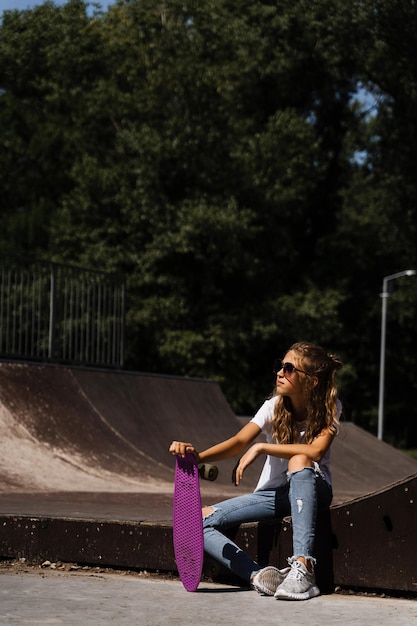 Enfant avec penny board Jeune fille adolescente dans des verres avec skate board assis sur une rampe de sport sur une aire de skate Mode de vie extrême Publicité créative pour skate ou penny board magasins ou magasins