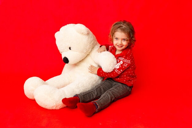 Enfant une petite fille avec un gros ours en peluche en vêtements d'hiver sur fond rouge, espace pour le texte