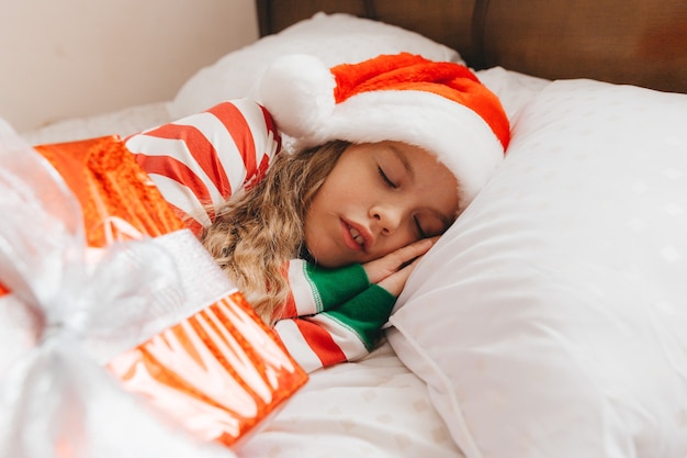 Un enfant, une petite fille en pyjama de Noël et chapeau de Père Noël, dort sur le lit à la maison.