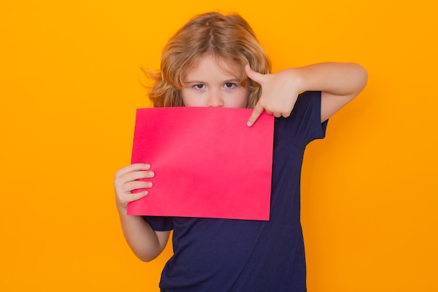 Enfant pointant sur une feuille de papier vide isolé sur fond jaune portrait d'un enfant tenant un bla