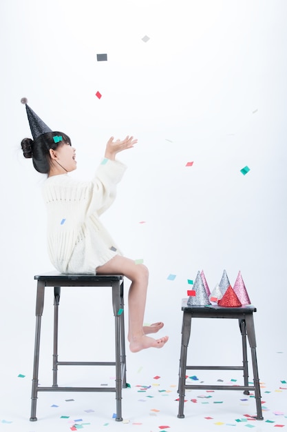 Un enfant portant un chapeau de fête noir est assis sur une chaise et s’amuse à regarder la poudre de papier colorée que sa mère jette.