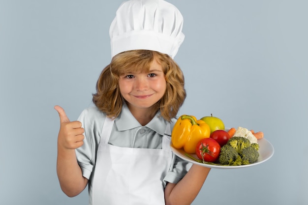 Enfant portant un uniforme de cuisinière et un chapeau de chef tenant une assiette avec des légumes préparant des légumes dans la cuisine