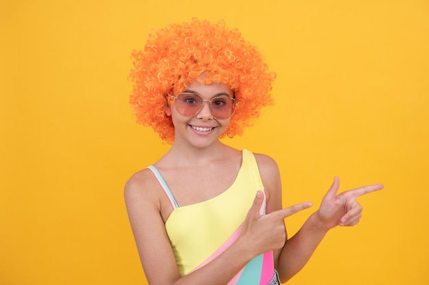 l'enfant porte des lunettes de soleil et un maillot de bain. adolescente aux cheveux orange pointant du doigt. mode de plage d'été. publicité. enfance heureuse. fête d'anniversaire. drôle d'enfant en perruque de clown frisée. s'amuser.