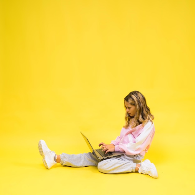 Enfant positif assis avec un ordinateur portable lire ses devoirs porter un tissu de style décontracté isolé sur fond jaune