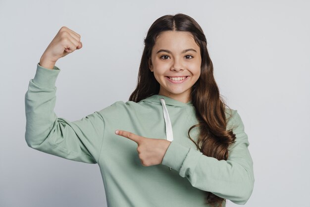 L'enfant positif pointe un doigt vers le biceps. Belle petite fille posant sur un fond de mur blanc