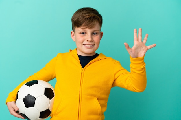 Enfant rousse jouant au football isolé sur fond bleu comptant cinq avec les doigts