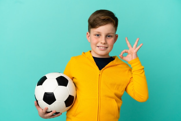 Enfant rousse jouant au football isolé sur fond bleu montrant un signe ok avec les doigts
