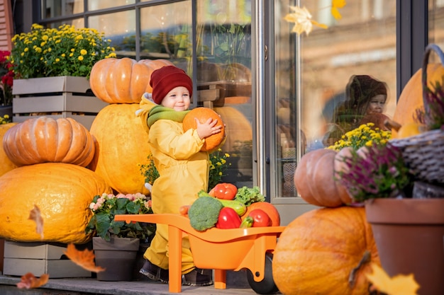 Un enfant en salopette jaune empile des légumes dans une petite voiture parmi de grosses citrouilles à la foire d'automne.