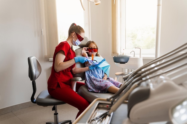 L'enfant se prépare à l'éclairage Uv du remplissage des dents par photopolymère en dentisterie Le dentiste dans des lunettes de protection rouges traite et élimine les caries chez une patiente enfant